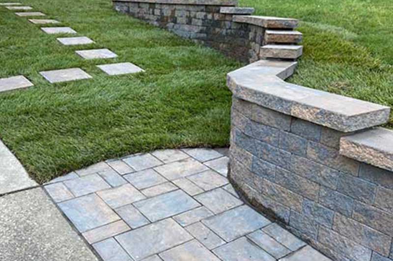 Grey brick walkway surrounded by grass and a grey wall in the green grass next to a spaced out light grey stone pathway
